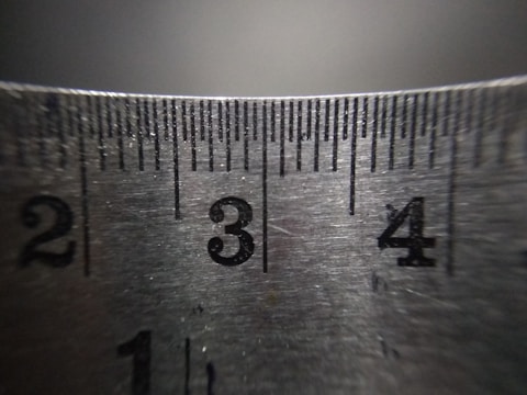 A close-up of a metallic ruler with engraved black numbers and lines indicating measurements. The surface reflects light subtly, showing its slightly worn texture.