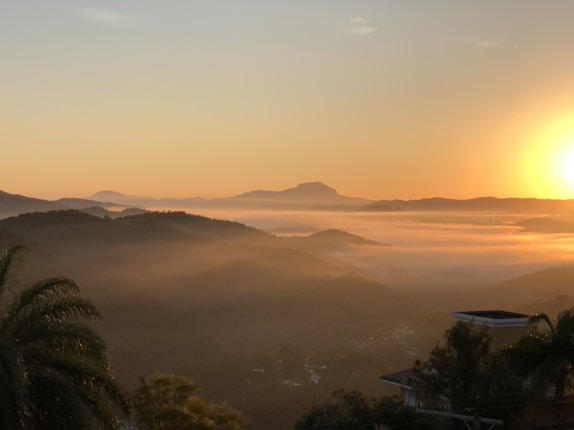 A serene landscape view of mountains covered with a layer of mist in the early morning. The sun rises on the right, casting a warm golden hue over the scene. A few trees and a building roof are visible in the foreground.
