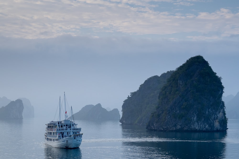 white ship on body of water near the rock formation covered with grass and tree