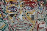 A vibrant mosaic artwork featuring a stylized human face. The face consists of small, colorful tiles arranged to form eyes, nose, and lips, with bold outlines in red. Various geometric shapes in yellow, green, blue, and white are used throughout the design to create a textured and patterned appearance.