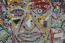 A vibrant mosaic artwork featuring a stylized human face. The face consists of small, colorful tiles arranged to form eyes, nose, and lips, with bold outlines in red. Various geometric shapes in yellow, green, blue, and white are used throughout the design to create a textured and patterned appearance.