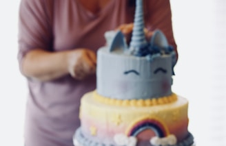 A person is decorating a two-tier cake with a unicorn theme. The top layer is blue with a unicorn horn and ears, featuring closed eyes drawn on. The bottom layer is decorated with a rainbow and small star shapes on a pastel gradient background.