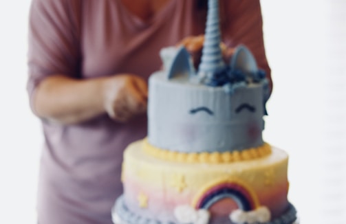 A person is decorating a two-tier cake with a unicorn theme. The top layer is blue with a unicorn horn and ears, featuring closed eyes drawn on. The bottom layer is decorated with a rainbow and small star shapes on a pastel gradient background.
