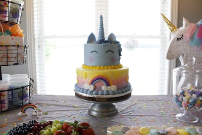 A whimsical unicorn-themed cake is centered on a decorative table. The cake features a blue and yellow ombre design with a unicorn horn and ears on top, with closed eyes as decoration. It is flanked by a colorful rainbow and flower accents. The table is covered with light purple cloth and scattered with colorful confetti. Nearby, there are baskets and containers with unicorn-themed party favors, along with an array of fruits like strawberries and grapes. To the side, a unicorn piñata adds to the festive atmosphere.