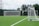 A well-maintained soccer field with lush green artificial turf is depicted. Two white soccer goals, one at a distance and another closer to the right, show the setup of the field. The goal nets are pristine and taut, indicating a space ready for a match or practice. Surrounding the field are tall trees providing a natural backdrop, and power lines are visible in the distance under a cloudy sky.