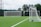 A well-maintained soccer field with lush green artificial turf is depicted. Two white soccer goals, one at a distance and another closer to the right, show the setup of the field. The goal nets are pristine and taut, indicating a space ready for a match or practice. Surrounding the field are tall trees providing a natural backdrop, and power lines are visible in the distance under a cloudy sky.