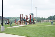 A playground with multiple slides and climbing structures made of red and green materials is positioned on a grassy field. In the background, there are sports fields with goal posts and a fence. Several people are visible, some walking and others participating in activities. Trees and a pavilion are also present in the park setting.