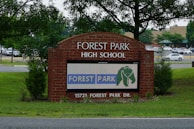 A brick sign for Forest Park High School is prominently displayed, surrounded by green grass and young trees. A digital display on the sign shows the text 'FOREST PARK' with an elephant logo. Behind the sign, a parking lot filled with cars and a background of more trees and a building are visible.
