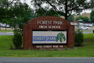 A brick sign for Forest Park High School is prominently displayed, surrounded by green grass and young trees. A digital display on the sign shows the text 'FOREST PARK' with an elephant logo. Behind the sign, a parking lot filled with cars and a background of more trees and a building are visible.