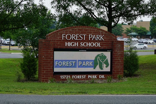 A brick sign for Forest Park High School is prominently displayed, surrounded by green grass and young trees. A digital display on the sign shows the text 'FOREST PARK' with an elephant logo. Behind the sign, a parking lot filled with cars and a background of more trees and a building are visible.