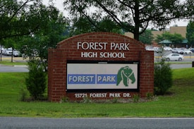 A brick sign for Forest Park High School is prominently displayed, surrounded by green grass and young trees. A digital display on the sign shows the text 'FOREST PARK' with an elephant logo. Behind the sign, a parking lot filled with cars and a background of more trees and a building are visible.