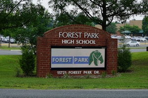 A brick sign for Forest Park High School is prominently displayed, surrounded by green grass and young trees. A digital display on the sign shows the text 'FOREST PARK' with an elephant logo. Behind the sign, a parking lot filled with cars and a background of more trees and a building are visible.