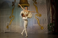 A ballet dancer is elegantly lifted by her partner on stage, surrounded by ornate gold and brown decorations resembling large bells. The dancers are dressed in traditional ballet attire, with the woman in a tutu and the man in tights. The backdrop depicts a village scene with buildings.