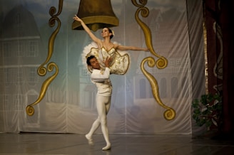 A ballet dancer is elegantly lifted by her partner on stage, surrounded by ornate gold and brown decorations resembling large bells. The dancers are dressed in traditional ballet attire, with the woman in a tutu and the man in tights. The backdrop depicts a village scene with buildings.
