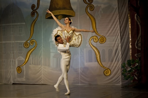 A ballet dancer is elegantly lifted by her partner on stage, surrounded by ornate gold and brown decorations resembling large bells. The dancers are dressed in traditional ballet attire, with the woman in a tutu and the man in tights. The backdrop depicts a village scene with buildings.