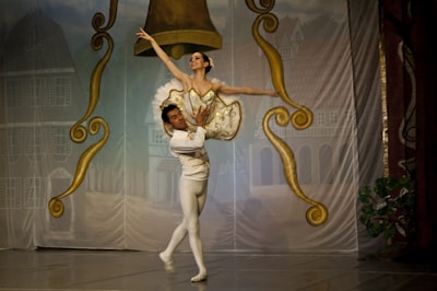 A ballet dancer is elegantly lifted by her partner on stage, surrounded by ornate gold and brown decorations resembling large bells. The dancers are dressed in traditional ballet attire, with the woman in a tutu and the man in tights. The backdrop depicts a village scene with buildings.