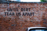 A brick wall has the phrase 'UNTIL DEBT TEAR US APART' spray-painted in black. A section of a parked car is visible at the bottom of the image, adding a touch of urban life.