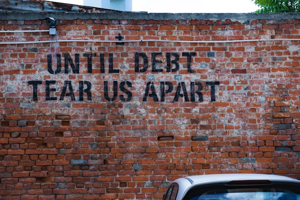 A brick wall has the phrase 'UNTIL DEBT TEAR US APART' spray-painted in black. A section of a parked car is visible at the bottom of the image, adding a touch of urban life.