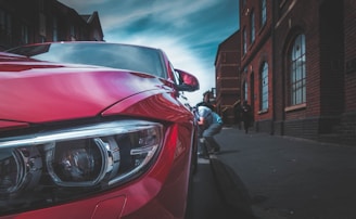 A red car is parked close to a curb on a street lined with brick buildings. One person is crouched down near the front of the car, possibly inspecting or fixing something. Another person is walking in the background, further down the sidewalk. The sky above is blue with some scattered clouds.