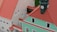A detailed model of a building complex, featuring red-tiled roofs and a prominent clock tower on a pastel green structure. The architecture includes multiple windows and entrance arches. The model is situated on a contrasting dark green background.