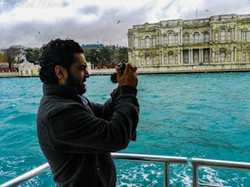 A person is taking a photograph on a boat with a historic building in the background across the water. The water is a vibrant turquoise, and the sky is cloudy with birds flying overhead.