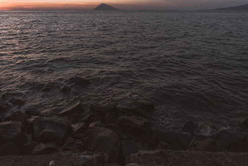 The image captures a tranquil coastal scene at dusk or dawn. The horizon features a distant silhouette of a mountain or large hill under a sky that is fading between shades of orange and gray. The foreground is dominated by the textured surface of the sea gently washing over dark, rugged rocks.