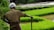 A farmer stands in a field of vibrant green rice paddies, holding a stick. The farmer is wearing a headscarf and has a muddy shirt, indicating recent work in the field.