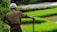 A farmer stands in a field of vibrant green rice paddies, holding a stick. The farmer is wearing a headscarf and has a muddy shirt, indicating recent work in the field.