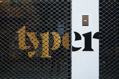 A metal security gate with a hexagonal pattern is placed in front of a building wall. Bright yellow and black letters are partially visible behind the gate. A white section of the wall features a black intercom panel near the top.
