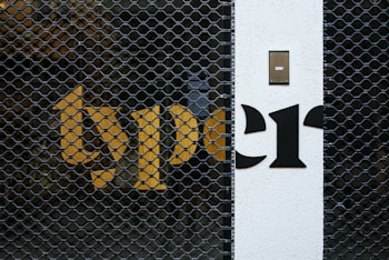 A metal security gate with a hexagonal pattern is placed in front of a building wall. Bright yellow and black letters are partially visible behind the gate. A white section of the wall features a black intercom panel near the top.