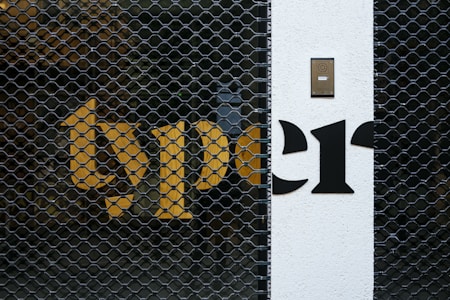 A metal security gate with a hexagonal pattern is placed in front of a building wall. Bright yellow and black letters are partially visible behind the gate. A white section of the wall features a black intercom panel near the top.