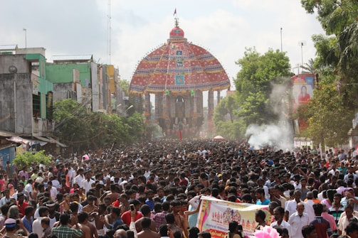 A large crowd of people gathers in a street participating in a colorful cultural or religious procession. A highly decorated temple chariot or rath is visible, adorned with intricate designs and vibrant colors. There are buildings lining the street and trees adding greenery. Smoke is emanating in certain areas, possibly from incense or ceremonial fires.
