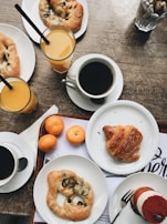 A breakfast setting includes a variety of pastries such as croissants and breads with olives on white plates, glasses of orange juice with straws, and cups of black coffee. There are also small oranges placed on the table. A piece of red and white layered cake is served on a small plate with a fork. The table surface is wooden, contributing to a cozy and rustic atmosphere.