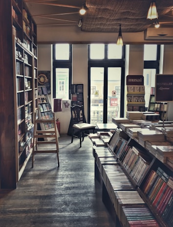 A cozy bookstore with wooden shelves on the left filled with books. A ladder is propped against the shelves, and a chair sits near the window. Stacks of books are also seen on the tables in the center of the room. Soft lighting from overhead lamps creates a warm ambiance.