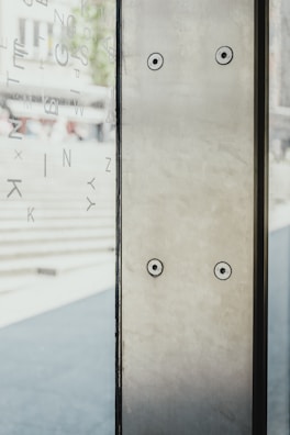 A glass panel stands vertically, featuring two round fasteners visible on the metallic surface. Letters are scattered on the glass, creating an abstract design. The background is blurred, showcasing a set of steps and indistinct structures.