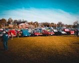 A gathering of classic cars is lined up on a grassy field with people walking around and inspecting the vehicles. Several brightly colored vintage cars and trucks are visible, with a backdrop of a small building, trees, and a clear blue sky.
