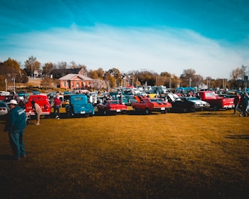 A gathering of classic cars is lined up on a grassy field with people walking around and inspecting the vehicles. Several brightly colored vintage cars and trucks are visible, with a backdrop of a small building, trees, and a clear blue sky.