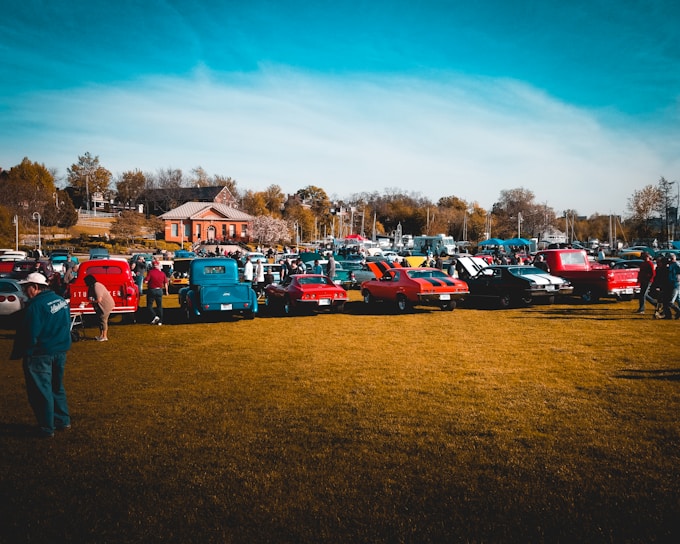 A gathering of classic cars is lined up on a grassy field with people walking around and inspecting the vehicles. Several brightly colored vintage cars and trucks are visible, with a backdrop of a small building, trees, and a clear blue sky.