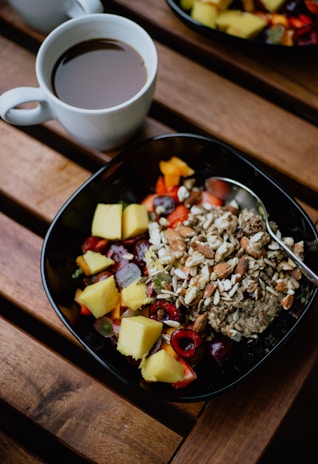 A cup of black coffee sits on a wooden table next to a bowl filled with colorful fruits, nuts, and grains. The bowl contains chunks of pineapple, strawberries, grapes, and assorted nuts and seeds, creating a vibrant and healthy breakfast scene.