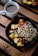 A cup of black coffee sits on a wooden table next to a bowl filled with colorful fruits, nuts, and grains. The bowl contains chunks of pineapple, strawberries, grapes, and assorted nuts and seeds, creating a vibrant and healthy breakfast scene.