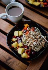 A cup of black coffee sits on a wooden table next to a bowl filled with colorful fruits, nuts, and grains. The bowl contains chunks of pineapple, strawberries, grapes, and assorted nuts and seeds, creating a vibrant and healthy breakfast scene.