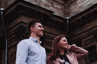 A smiling young man and woman stand closely together, gazing upwards in front of a large, ornate stone building. The man is wearing a light blue shirt, while the woman is dressed in a pink jacket. The mood is cheerful and relaxed, with a sense of admiration for their surroundings.
