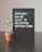 A black pegboard sign displaying the motivational quote 'DIFFICULT ROADS LEAD TO BEAUTIFUL DESTINATIONS' in white lettering. Next to it, a small potted plant with green leaves rests on a dark surface, likely a table. The background features a neutral, light-colored wall.