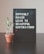 A black pegboard sign displaying the motivational quote 'DIFFICULT ROADS LEAD TO BEAUTIFUL DESTINATIONS' in white lettering. Next to it, a small potted plant with green leaves rests on a dark surface, likely a table. The background features a neutral, light-colored wall.