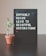 A black pegboard sign displaying the motivational quote 'DIFFICULT ROADS LEAD TO BEAUTIFUL DESTINATIONS' in white lettering. Next to it, a small potted plant with green leaves rests on a dark surface, likely a table. The background features a neutral, light-colored wall.
