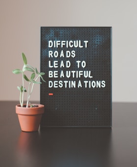 A black pegboard sign displaying the motivational quote 'DIFFICULT ROADS LEAD TO BEAUTIFUL DESTINATIONS' in white lettering. Next to it, a small potted plant with green leaves rests on a dark surface, likely a table. The background features a neutral, light-colored wall.