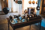A cozy kitchen scene featuring an assortment of vintage kitchenware. The central focus is a dark wooden table with a silver tray holding blue and white porcelain teacups and a milk jug. To the right, a dark brown tray holds various copper pots and kettles. Behind the table, large copper pots and pans hang on the stone walls, contributing to the rustic and antique aesthetic. A large, dark ceramic pot is visible to the left, adding to the traditional charm of the room.