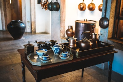 A cozy kitchen scene featuring an assortment of vintage kitchenware. The central focus is a dark wooden table with a silver tray holding blue and white porcelain teacups and a milk jug. To the right, a dark brown tray holds various copper pots and kettles. Behind the table, large copper pots and pans hang on the stone walls, contributing to the rustic and antique aesthetic. A large, dark ceramic pot is visible to the left, adding to the traditional charm of the room.