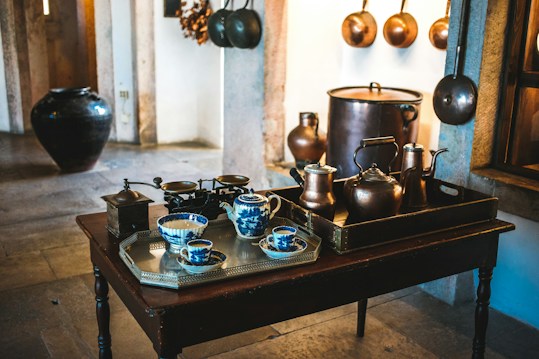 A cozy kitchen scene featuring an assortment of vintage kitchenware. The central focus is a dark wooden table with a silver tray holding blue and white porcelain teacups and a milk jug. To the right, a dark brown tray holds various copper pots and kettles. Behind the table, large copper pots and pans hang on the stone walls, contributing to the rustic and antique aesthetic. A large, dark ceramic pot is visible to the left, adding to the traditional charm of the room.