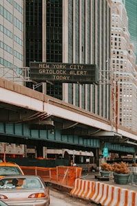 An urban scene featuring tall buildings with a digital traffic sign displaying a gridlock alert. A roadway is partially visible below with construction barriers and cars. High-rise structures dominate the background, emphasizing a city environment.